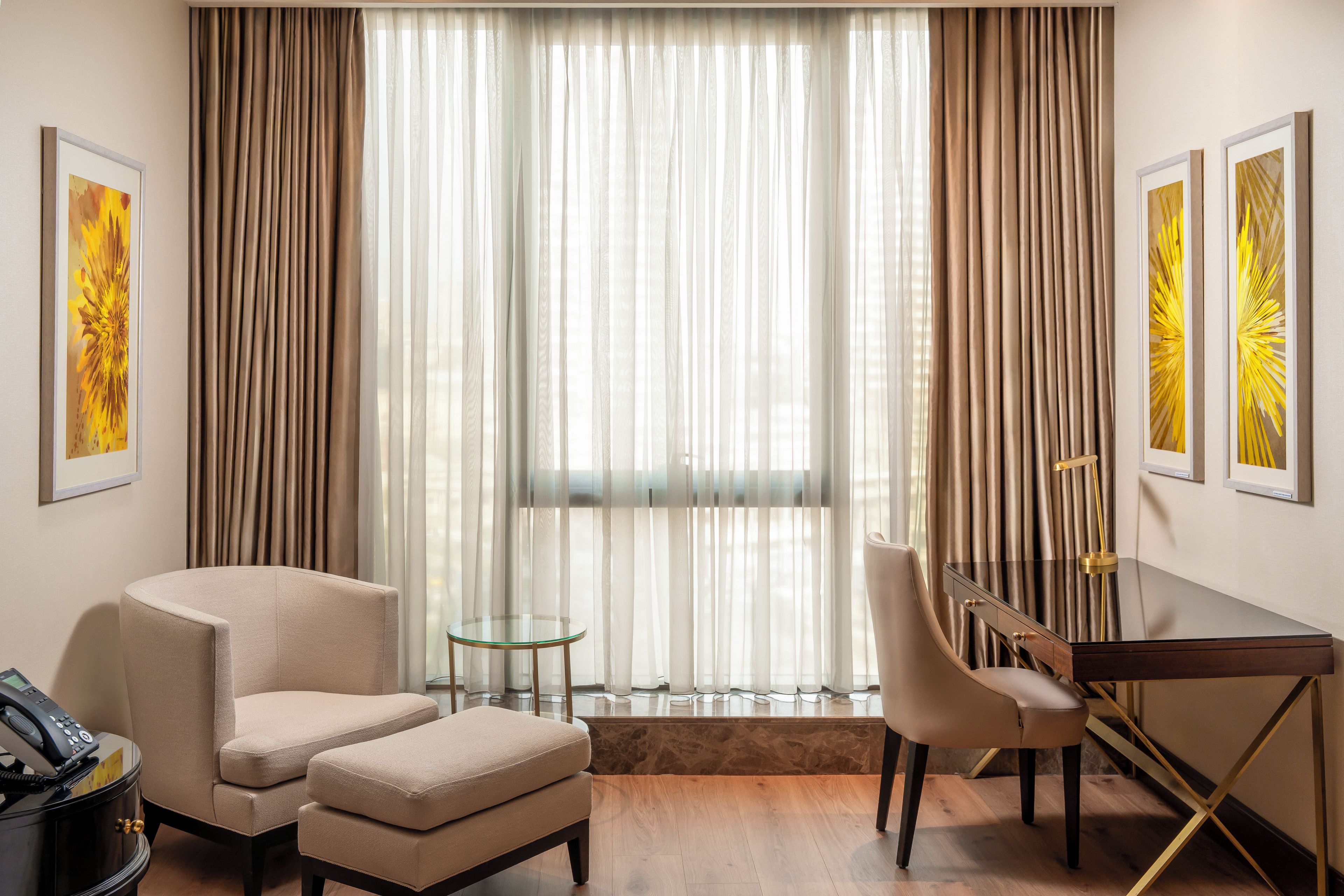 A hotel room with a large window, a desk, two chairs, a small round table, and artwork on the walls.