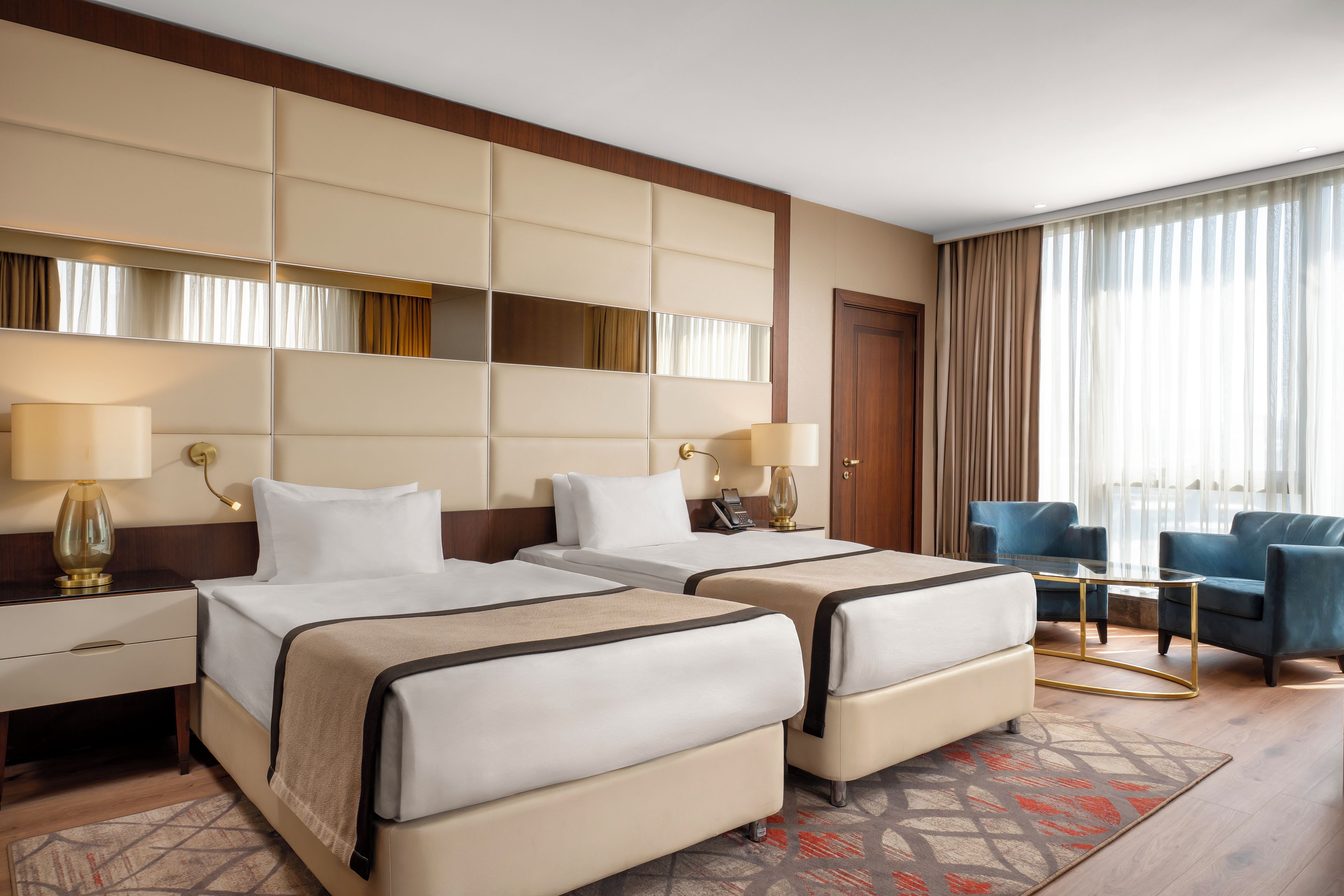 A hotel room with two beds, a desk, a chair, and a large window with curtains.