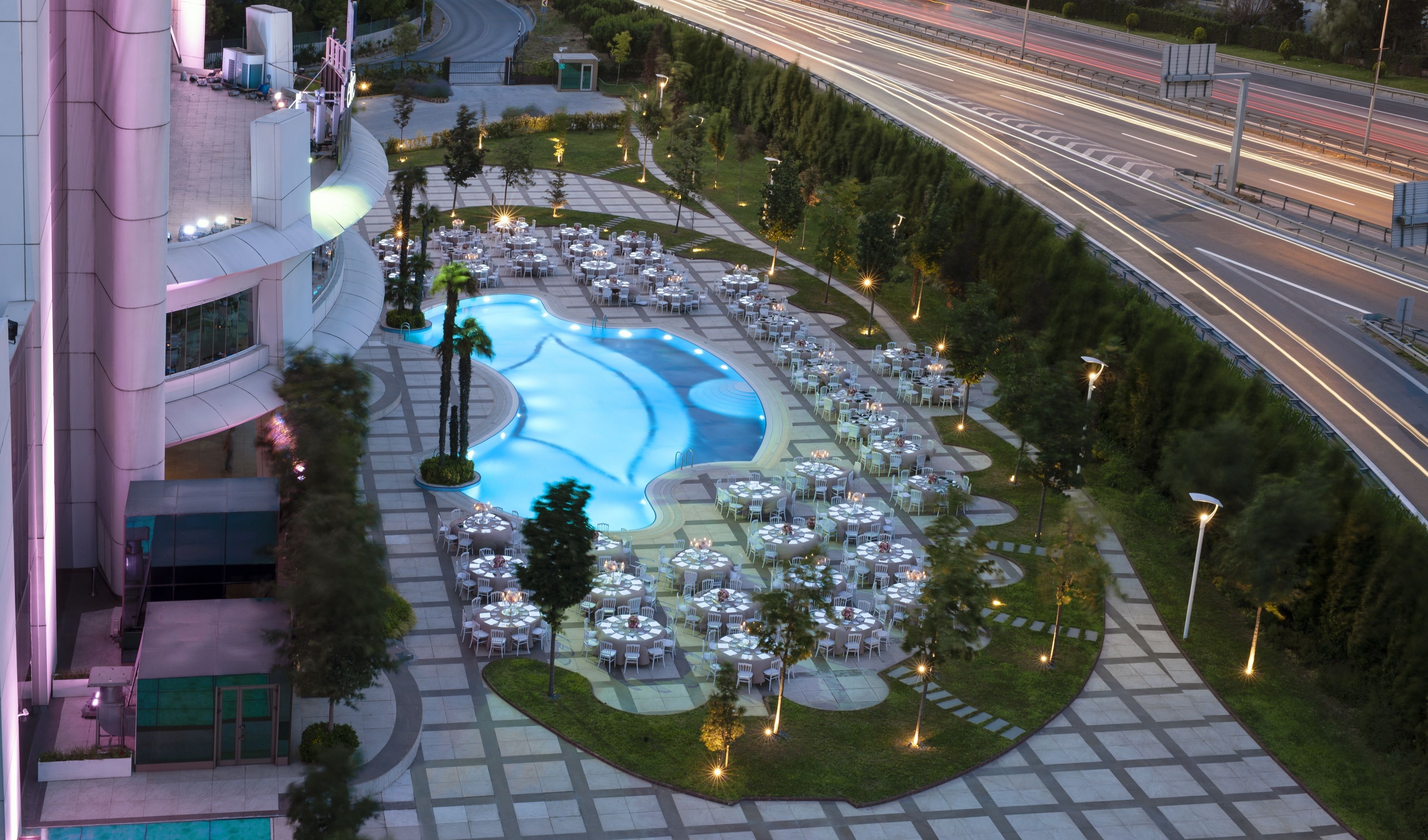 An outdoor pool area with multiple round tables and chairs, surrounded by greenery and pathways.