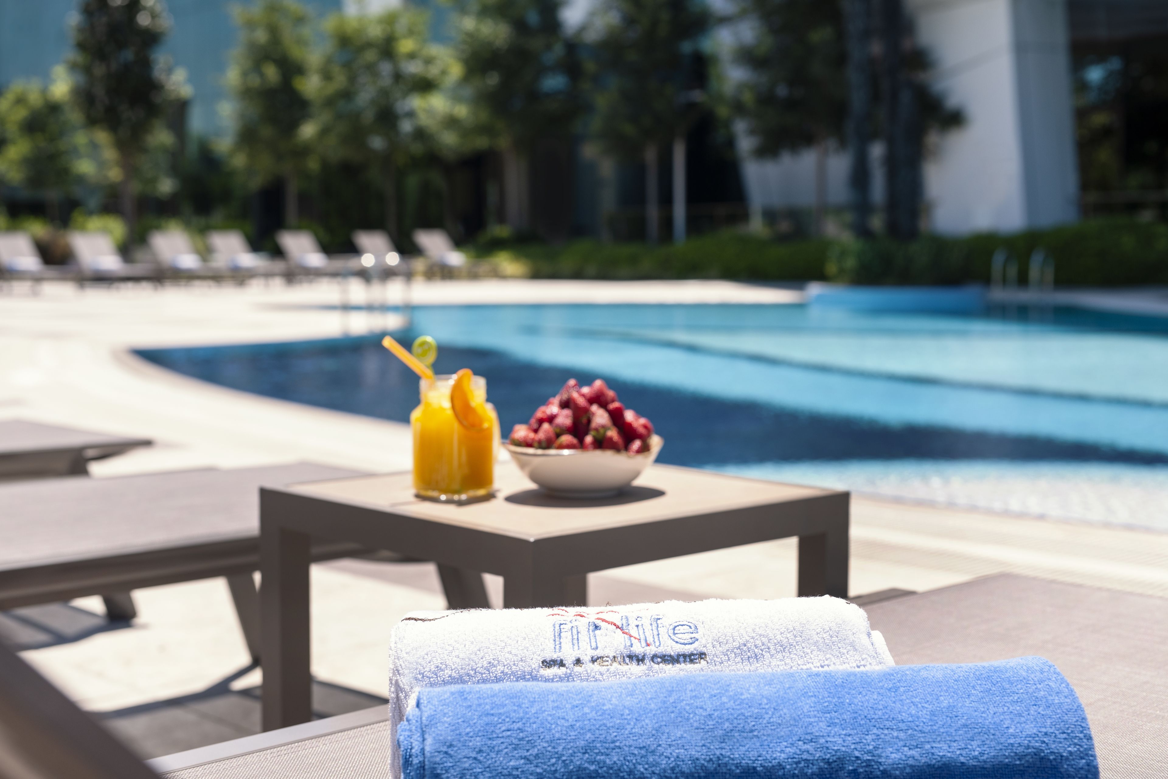 Zona junto a la piscina con sillones, una mesa con un frutero y una bebida, y una toalla con el nombre del hotel.