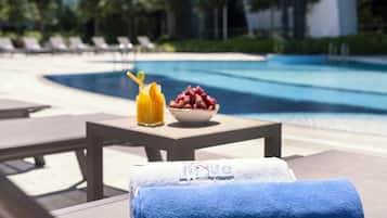 Area piscina con lettini, un tavolo con una ciotola di frutta e bevande, e un asciugamano con il nome dell'hotel.
