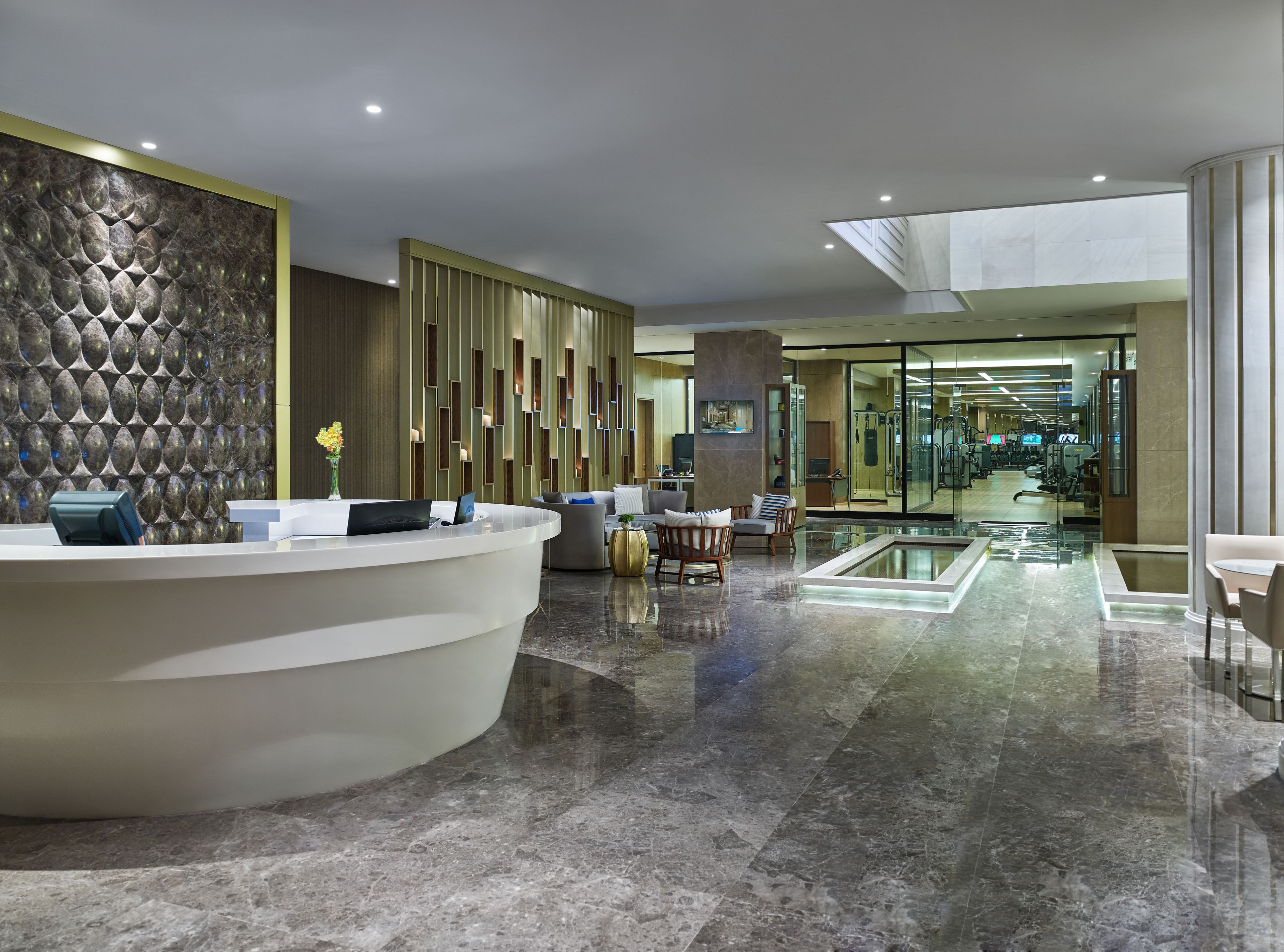 Een moderne hotellobby met een receptiebalie, een zithoek en een fitnessruimte met glazen wanden.