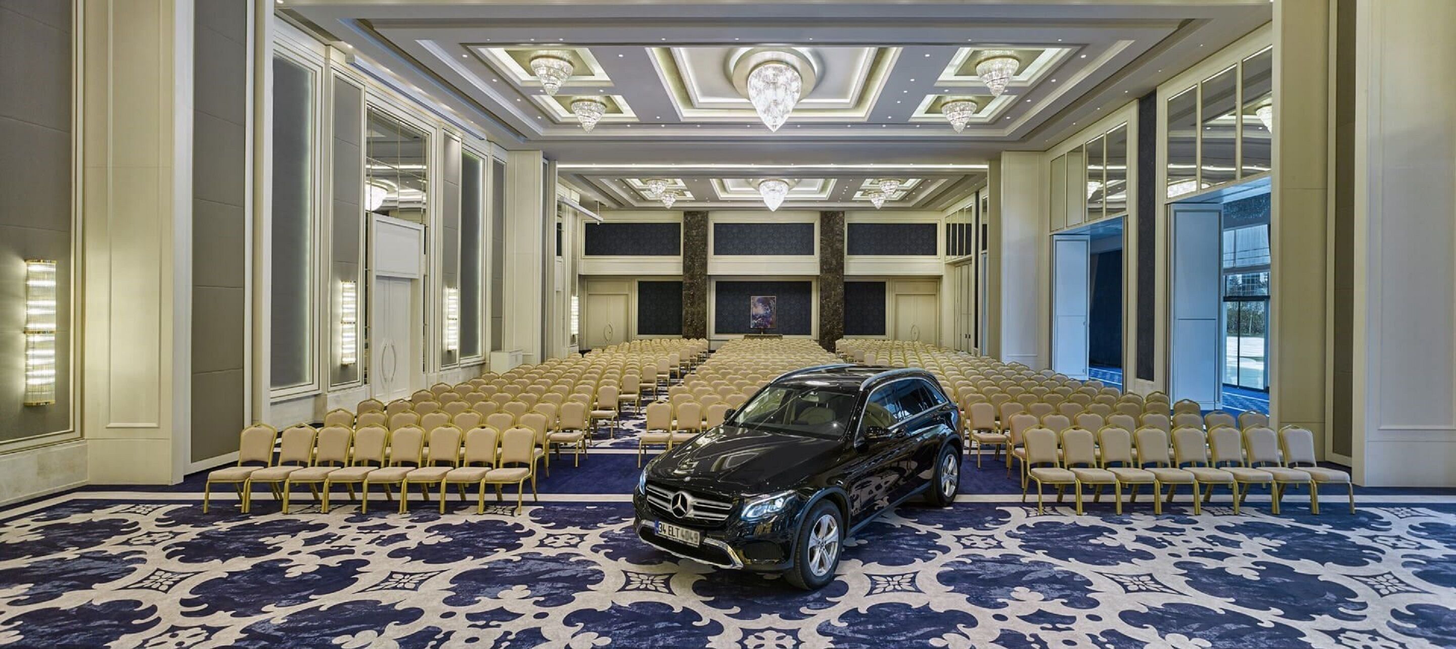 A spacious conference room with rows of chairs, a black car parked in the center, and a patterned carpet.