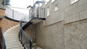 Un escalier avec une rampe en métal noir mène à un palier. Les murs sont recouverts de carreaux de pierre, et un panneau en bois se trouve sur le côté gauche.