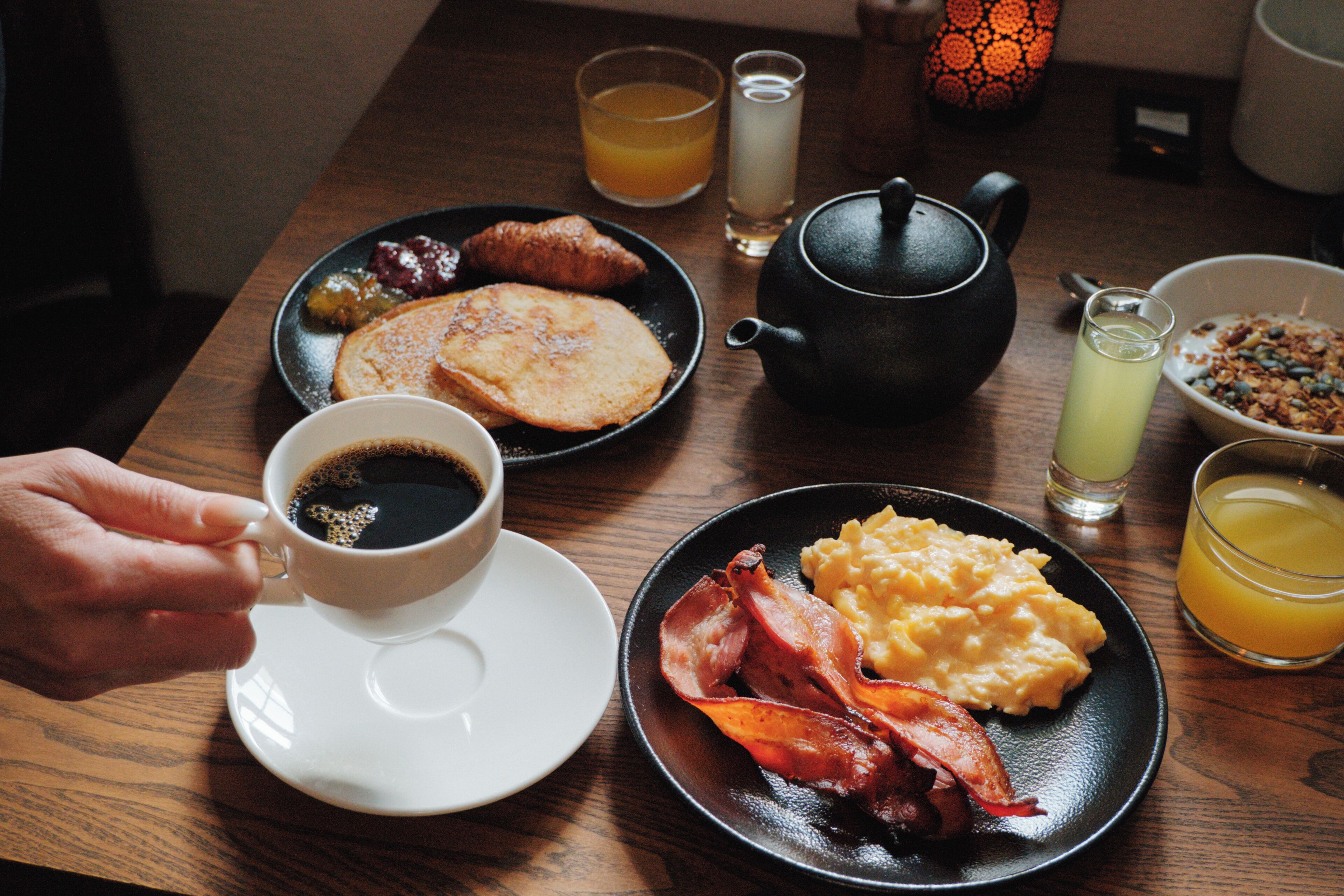 Una mesa de desayuno con platos de comida, un calentador de té, tazas y vasos de jugo de naranja.
