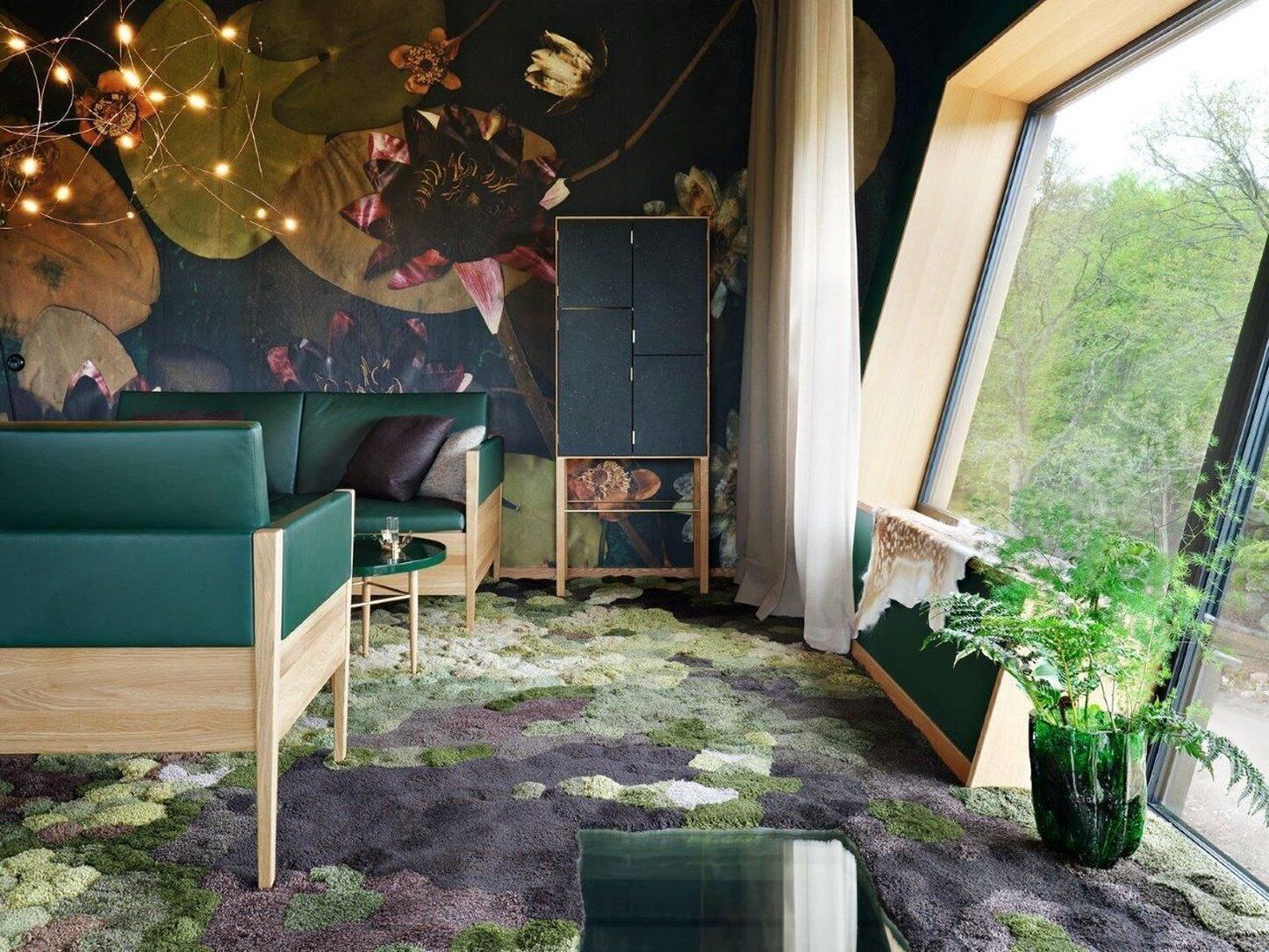 Ein gemütliches Zimmer mit einem grünen Sofa, einem kleinen Tisch und einem großen Fenster mit Blick ins Grüne.