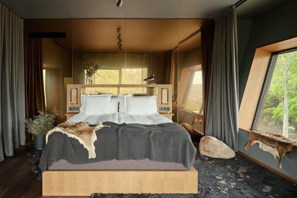 A bedroom with a large bed, a wooden headboard, a window with a view of trees, and a stone wall.