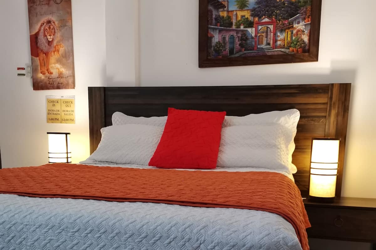 Un lit bien fait, recouvert d’une couvre-lit blanche et orange, avec deux oreillers blancs et un oreiller décoratif rouge. Deux lampes de chevet sont placées de chaque côté du lit.