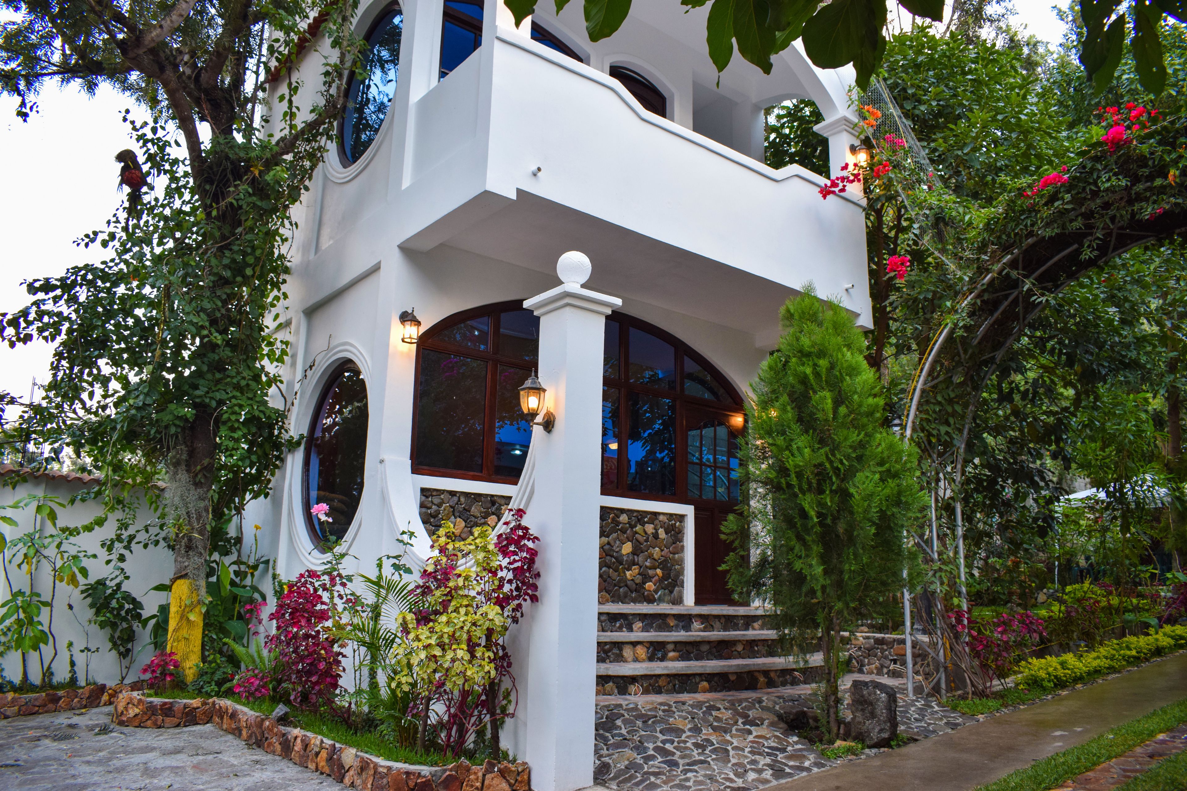 Um edifício de dois andares com varanda, degraus de pedra e um jardim com diversas plantas e flores.