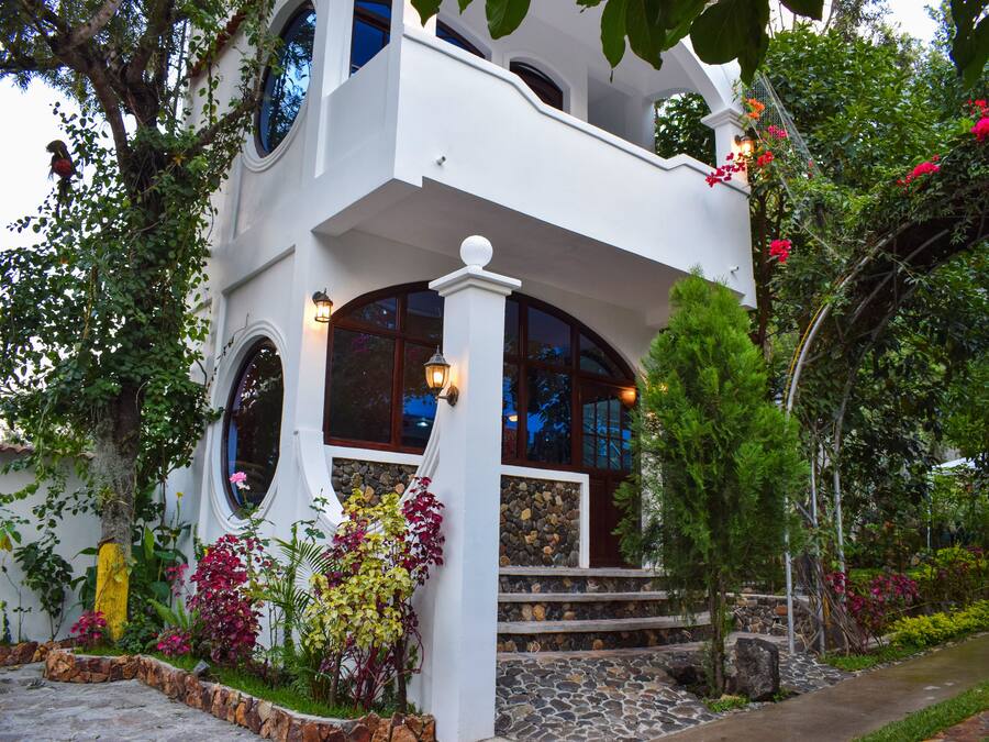 Um edifício de dois andares com varanda, degraus de pedra e um jardim com diversas plantas e flores.
