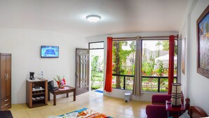 Sala de estar con televisor, muebles de madera, una mesita con una computadora portátil y vistas a un jardín a través de amplias ventanas.