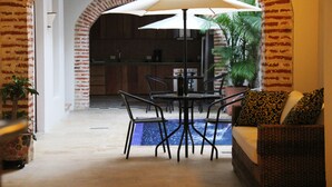 An indoor patio area with a table and chairs under a large umbrella, a sofa with patterned cushions, and a view of a kitchen through an arched doorway.
