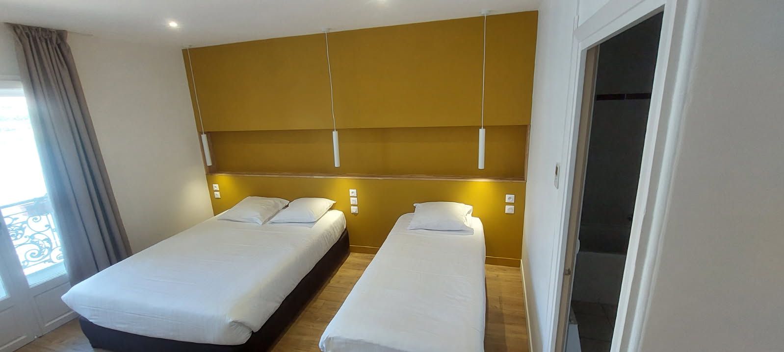 Quarto de hotel com duas camas, piso de madeira, parede amarela com detalhes e uma varanda com vista.