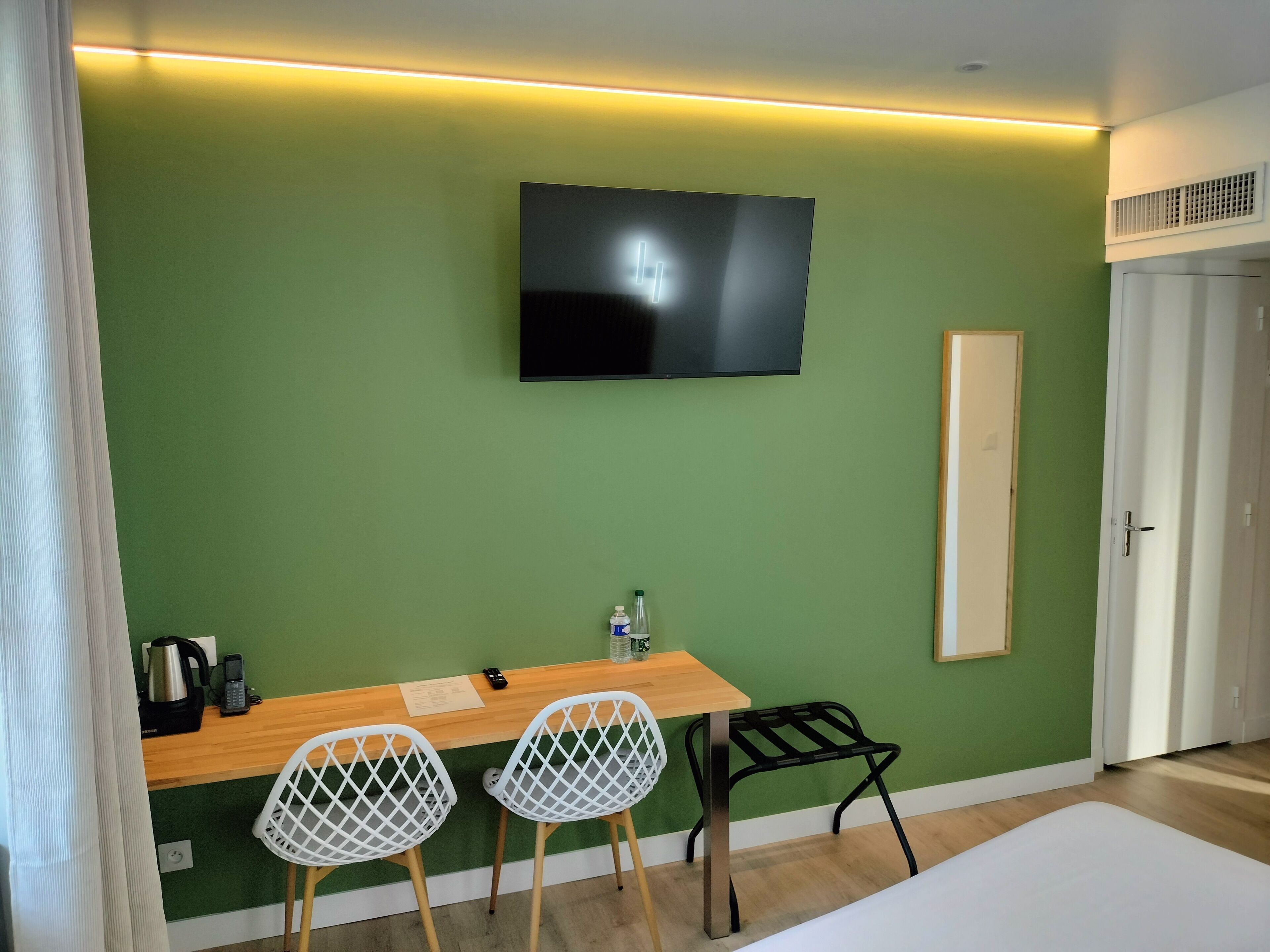 Une pièce avec un mur vert, un téléviseur à écran plat fixé au-dessus d’une table en bois, deux chaises blanches, une petite lampe de table et une bouteille d’eau.