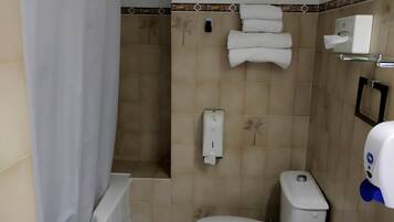 A small bathroom with a toilet, a sink, and a shower. There are towels hanging on the wall and a soap dispenser.