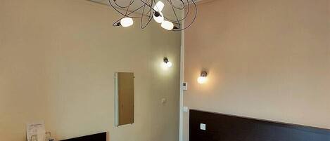 A hotel room with a bed, a nightstand, a minibar, a mirror, and a modern chandelier.