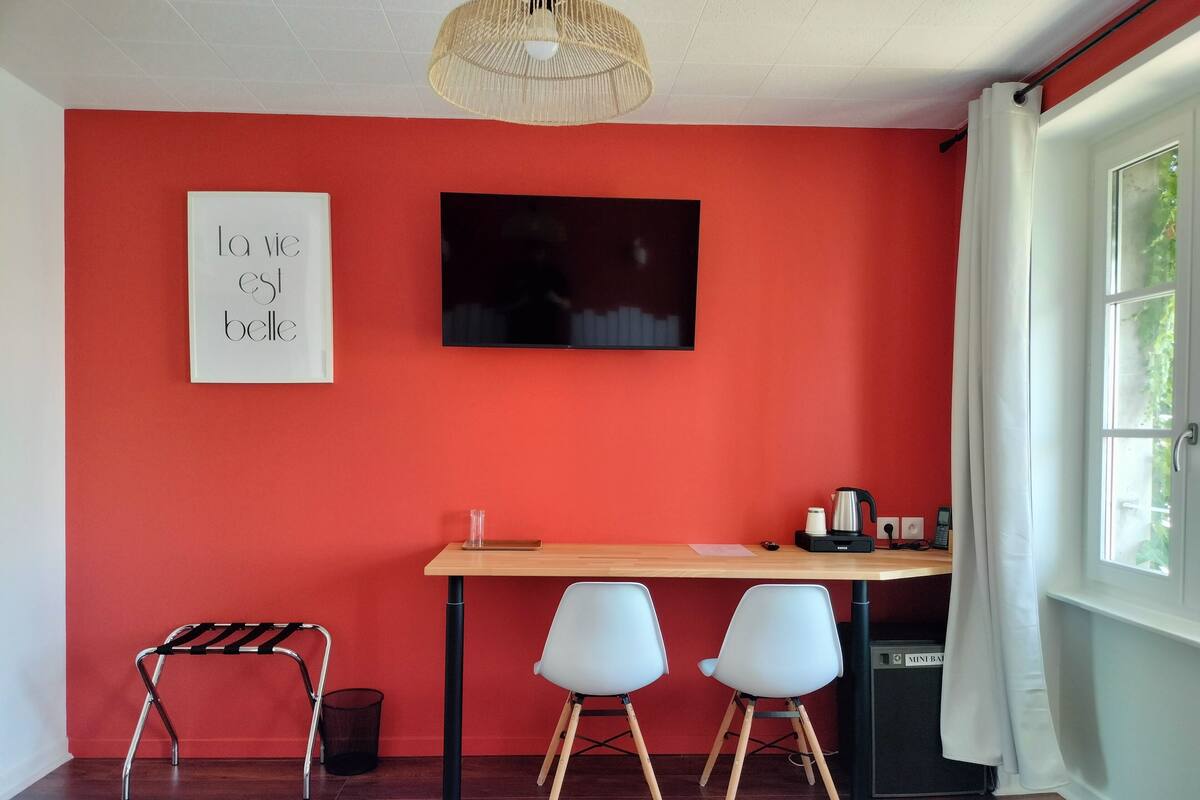 A room with a red wall, a wooden desk, two white chairs, a black TV, a coffee maker, and a wicker lamp.