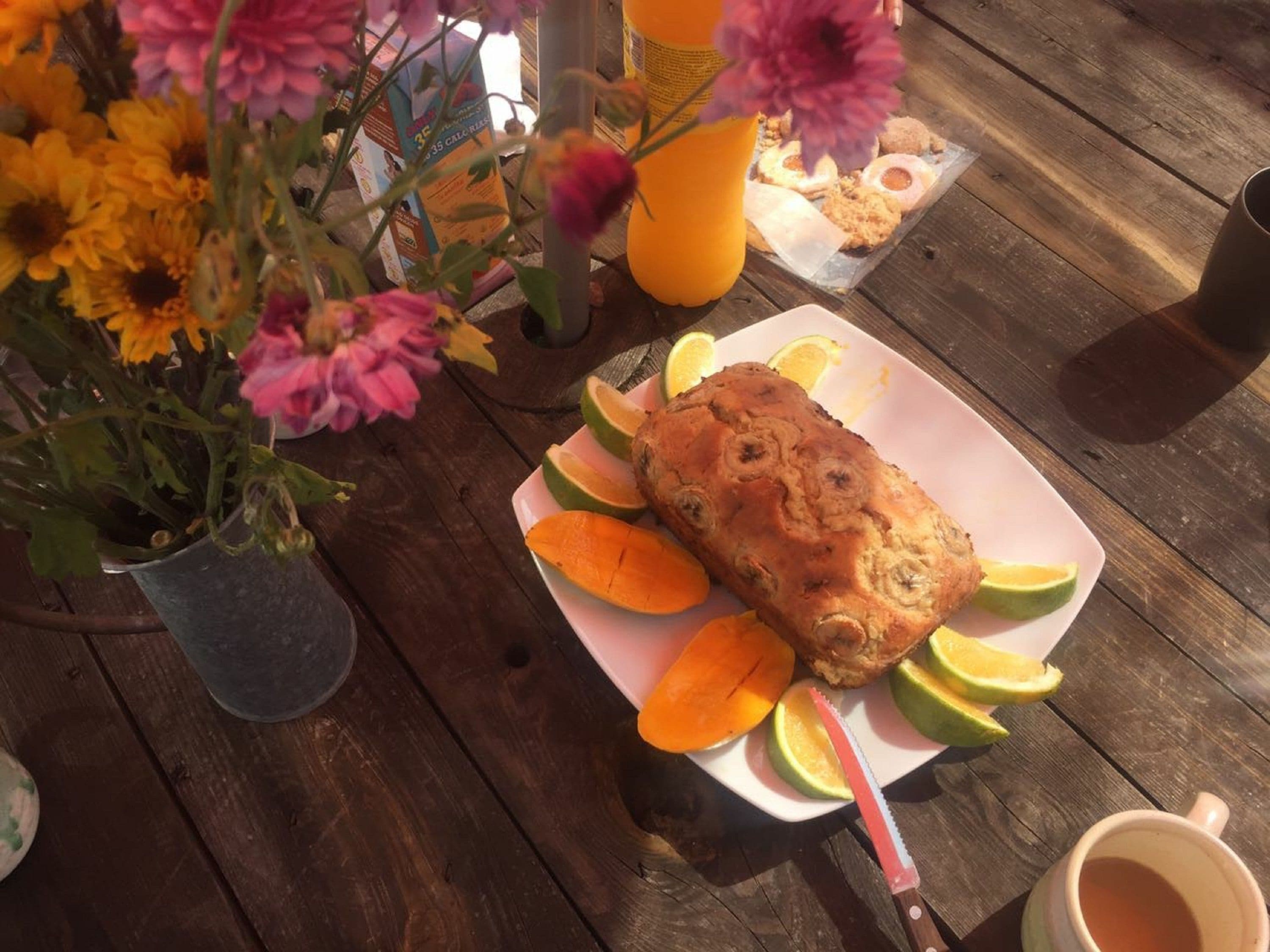 Une miche de pain sur une assiette, accompagnée de tranches de citron vert et d’orange, d’un couteau et d’une tasse de thé, le tout posé sur une table en bois.