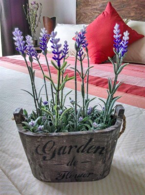 Un bac à fleurs en bois rustique, contenant des plants de lavande, est posé sur un lit agrémenté d’un coussin rouge et, en arrière-plan, d’un coussin blanc.