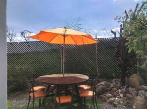 Um pátio externo com mesa redonda de madeira e cadeiras laranjas, um guarda-sol laranja e uma cerca de arame atrás.