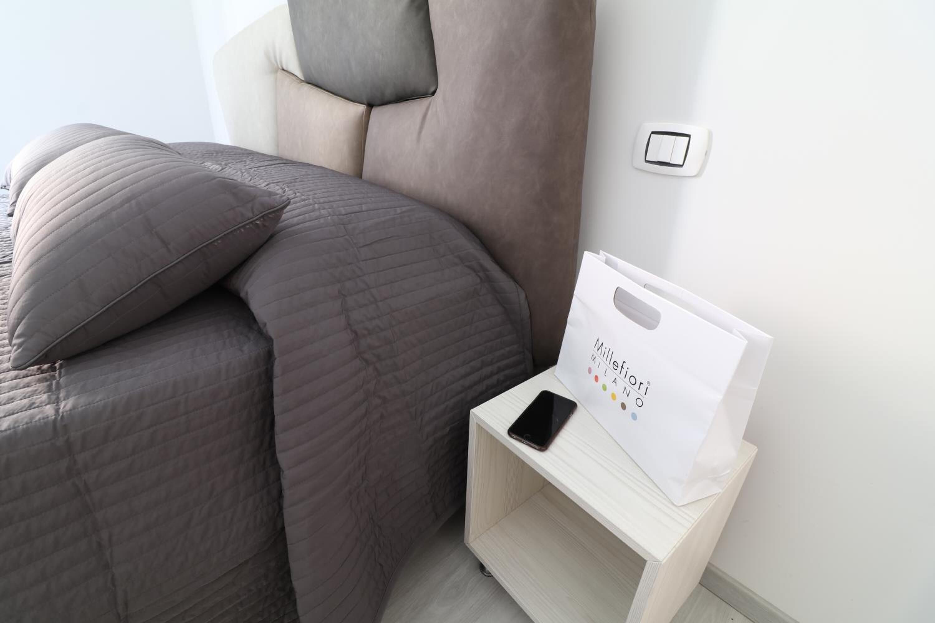 Una cama bien tendida con un edredón grisáceo y almohadas, una mesita de noche con un bolso blanco y un teléfono inteligente, y un interruptor de luz instalado en la pared.