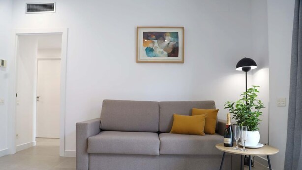 A modern living room with a sofa, a small table with a plant and a bottle, and a framed artwork on the wall. - Cosmo Apartments Concell de Cent (Barcelona)