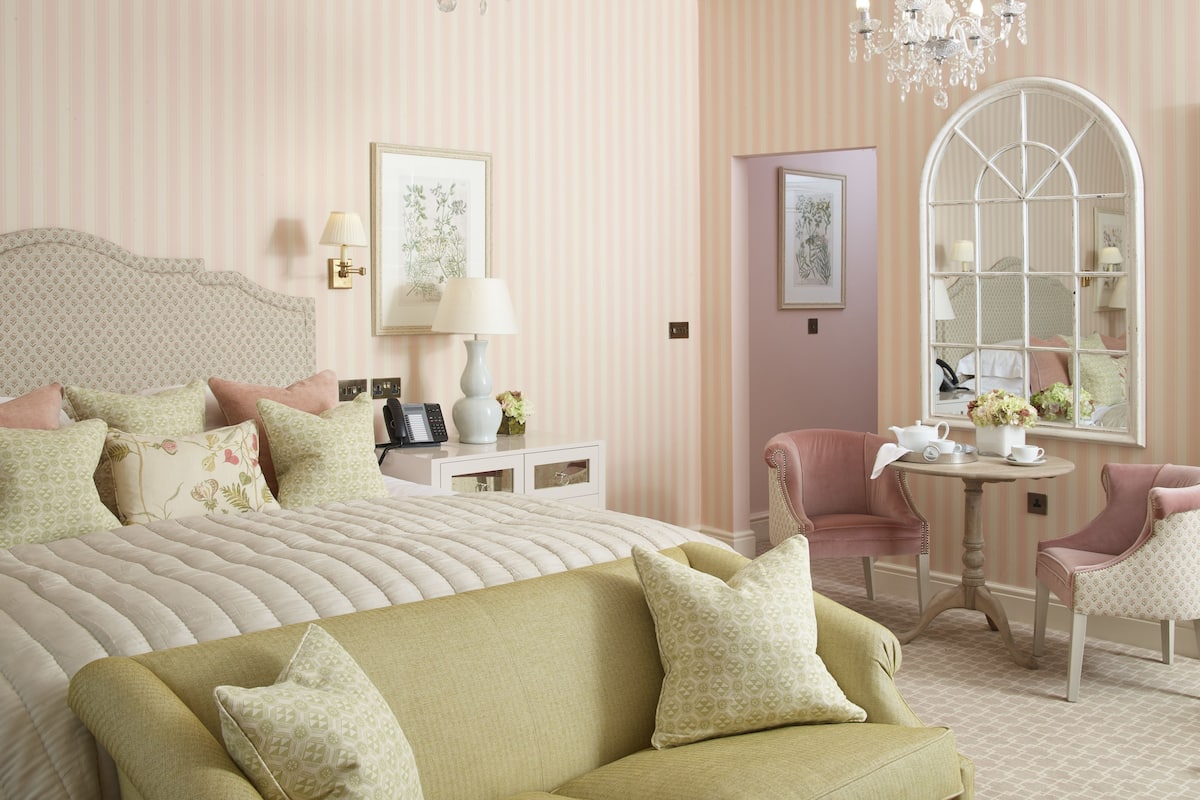 Una habitación de hotel con una cama grande, una mesita de noche con lámpara, una silla, una pequeña mesa con un florero y un espejo.