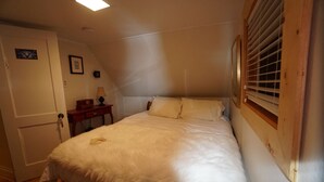 A bedroom with a bed, a door, a small table with a lamp, and a window with blinds. - White On Wood (Crow Head)