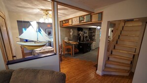 A living room with a wooden floor, a sofa, and a sailboat model. A staircase leads to a kitchen area with a wooden counter and appliances. - White On Wood (Crow Head)