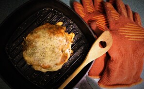 A grilled chicken breast on a grill pan with a wooden spatula and orange oven mitts beside it. - White On Wood (Crow Head)