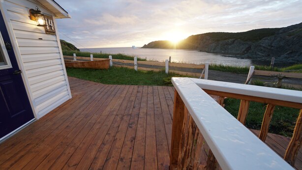 A wooden deck with a railing, a door, and a view of the ocean at sunset. - White On Wood (Crow Head)