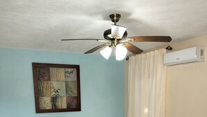 Habitación con dos camas, ventilador de techo, aire acondicionado, una mesita pequeña y un adorno en la pared.
