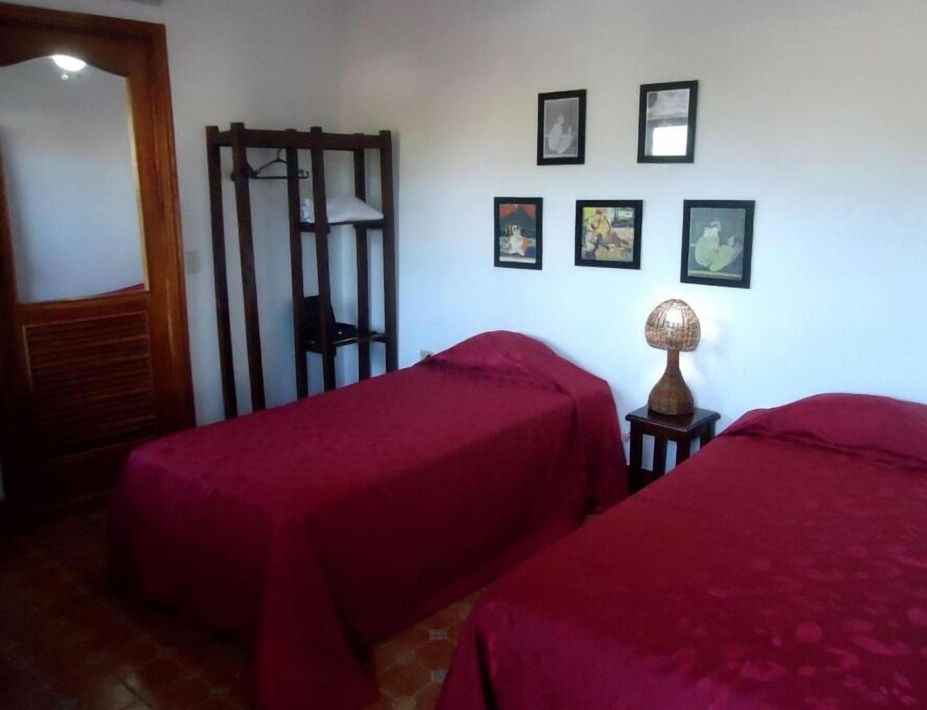 Une pièce avec deux lits recouverts de draps rouges, une chaise en bois avec un dossier, une petite table avec une lampe et quatre tableaux encadrés accrochés au mur.
