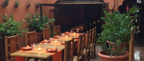 An outdoor dining area with wooden tables and chairs, potted plants, and a tiled floor.