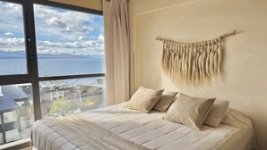 Un dormitorio con un ventanal grande, una cama con ropa de cama color beige y un tapiz de macramé en la pared.
