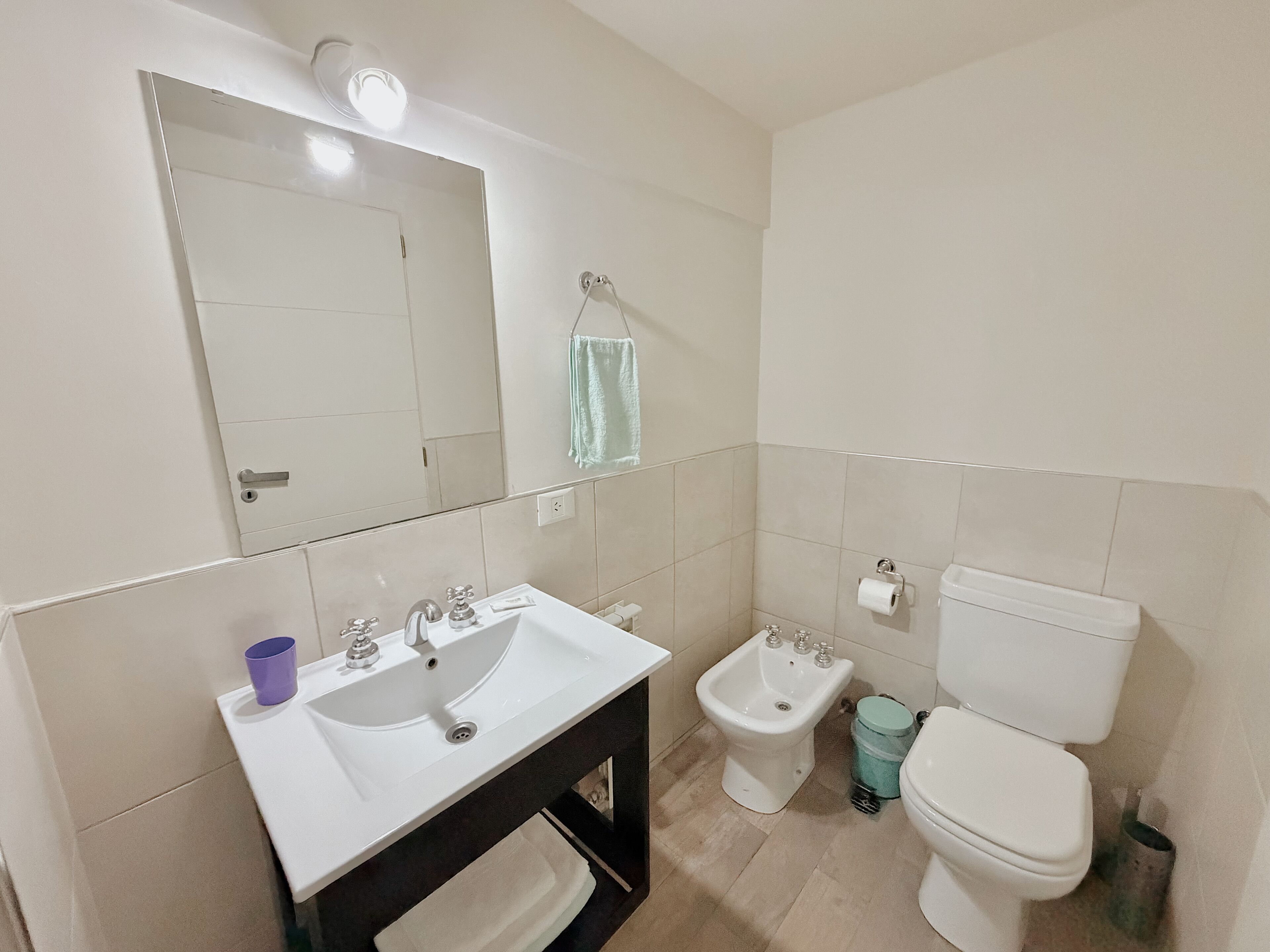 A bathroom with a sink, mirror, bidet, toilet, and a trash bin.