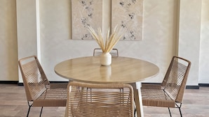 Una mesa redonda de madera con una silla tejida a cada lado, un florero con plantas secas y dos cuadros abstractos en la pared.