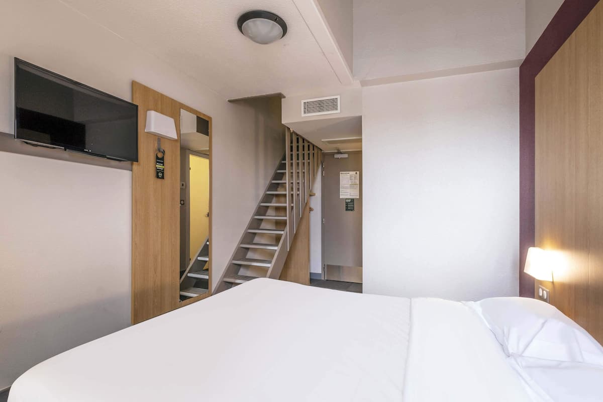 A hotel room with a bed, a TV mounted on the wall, a staircase leading to another room, and a wall-mounted light fixture.