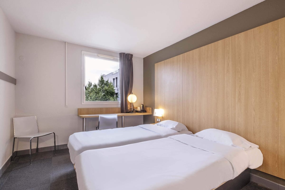 A hotel room with two beds, a desk, a chair, and a window with a view of greenery.