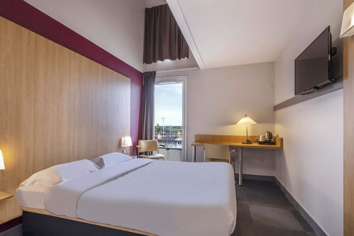 A hotel room with a large bed, a desk, a chair, a TV, and a window with a city view.