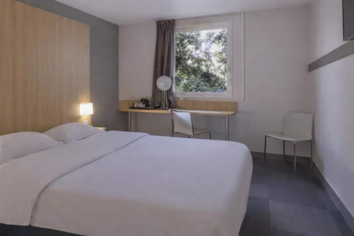 A hotel room with a bed, a desk, a chair, a window, and a lamp.