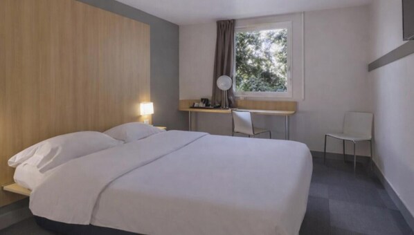 A hotel room with a bed, a desk, a chair, a window, and a lamp.
