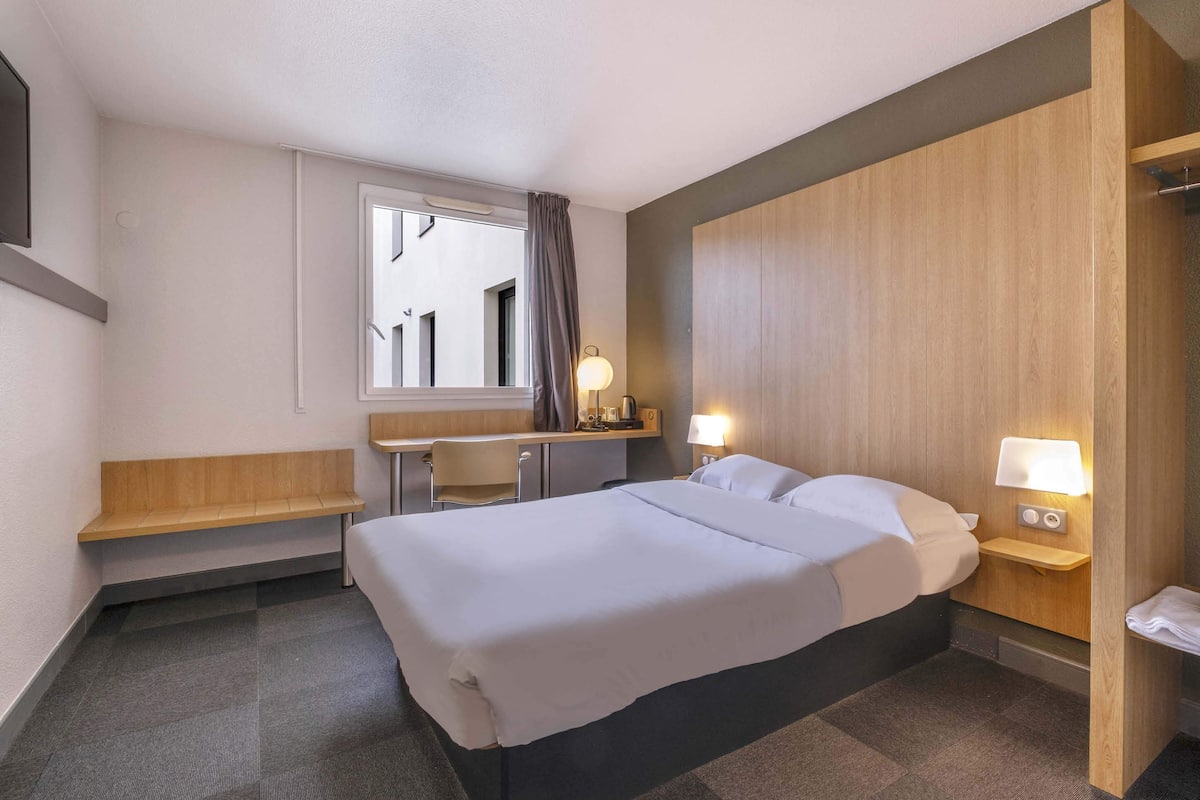 A hotel room with a bed, a desk, a chair, a small table, a nightstand with a lamp, and a window with curtains.