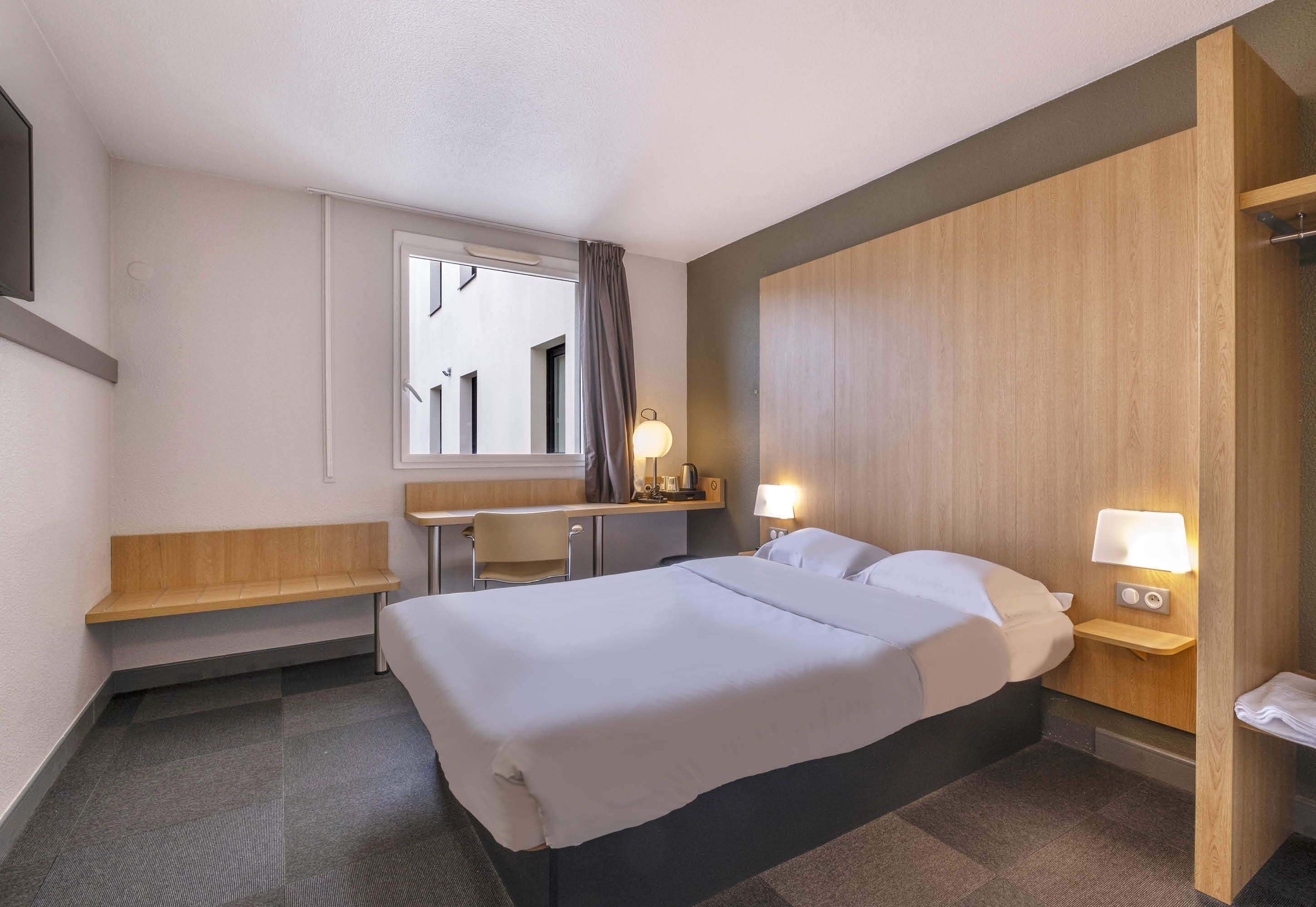 Una habitación de hotel con cama, escritorio, silla, una mesita, lámpara y ventana con cortinas.