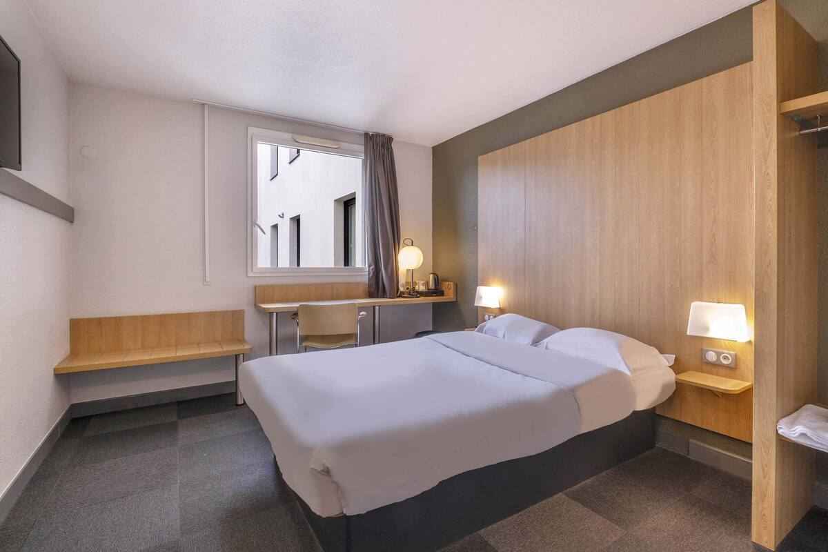 A hotel room with a bed, a desk, a chair, a small table, a nightstand with a lamp, and a window with curtains.