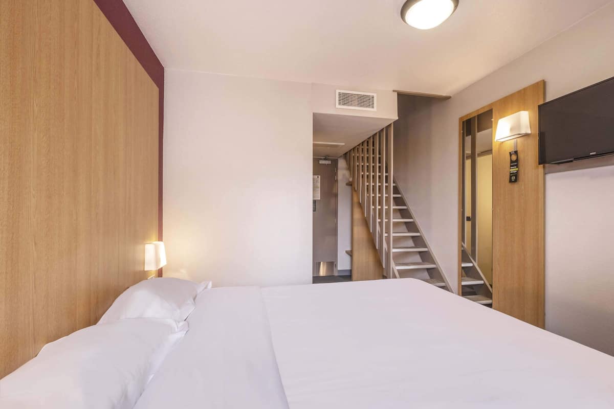 A hotel room with a bed, a wooden headboard, a TV mounted on the wall, and a staircase leading to another room.