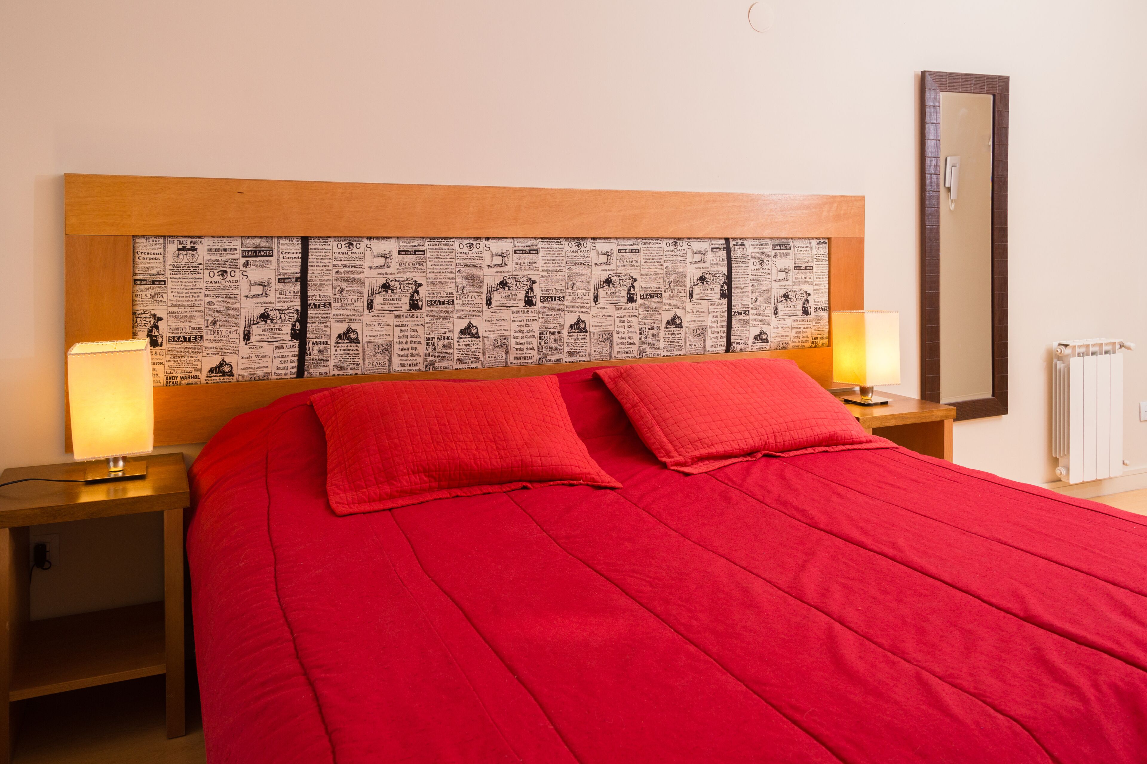 Una camera d'albergo con una coperta rossa, due cuscini, una testiera di legno con stampa a righe di giornale, due lampade ai comodini e uno specchio sulla parete.