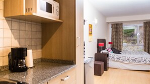 Eine kompakte Küche mit Mikrowelle, Kaffeemaschine und kleinem Kühlschrank. Angrenzend befindet sich ein Schlafzimmer mit Bett, Nachttisch und einem Vorhangfenster.