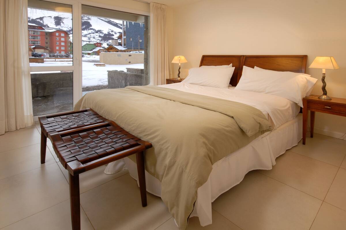 Habitación de hotel con cama, dos mesitas de noche con lámparas, un banco de madera y vistas a una montaña nevada y edificios.