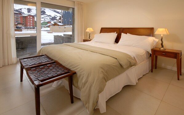 A hotel room with a bed, two bedside tables with lamps, a wooden bench, and a view of a snowy mountain and buildings. - Catedral Apartments (San Carlos de Bariloche)